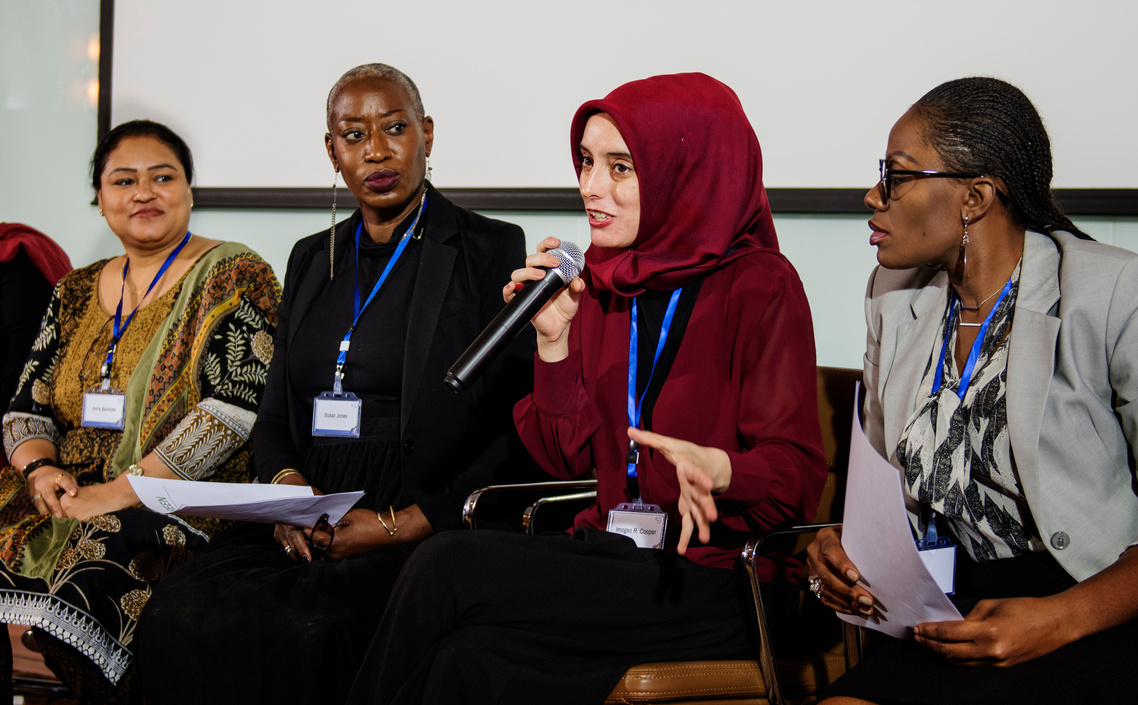 Photo of a group of diverse women at a conference, with one speaking into a microphone