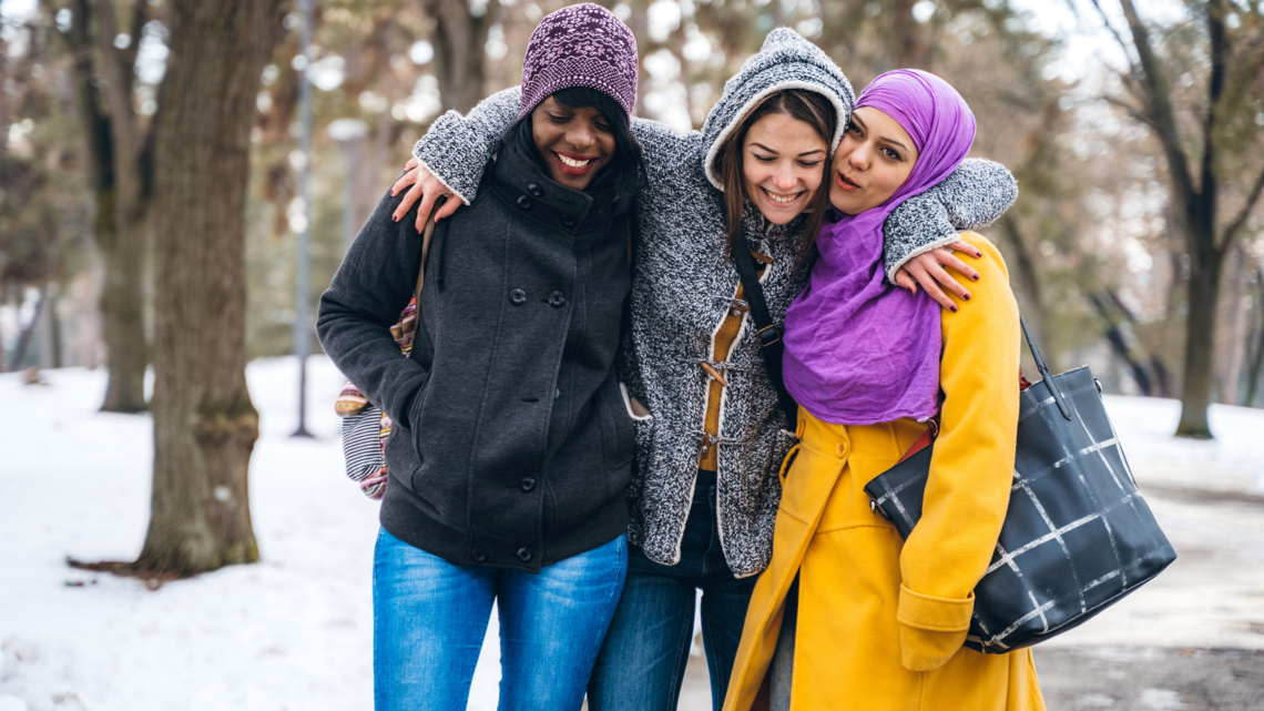 A photo of three women walking with arms around each other and smiling in a snowy woodland