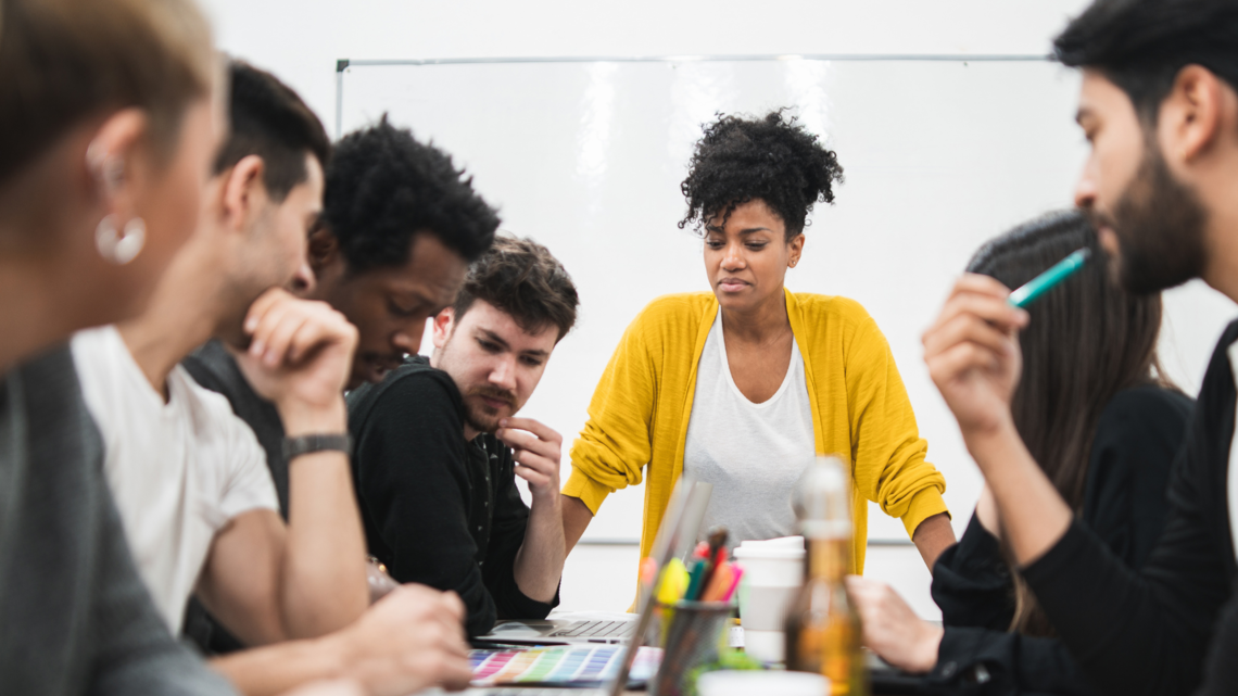 Photo of a diverse team sat working at a table with a woman stood up talking